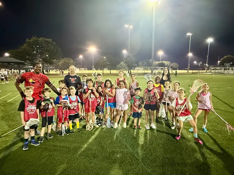 Boys and girls from the Parkland Redhawks smile with their lacrosse sticks, with two coaches, on the field at a night session